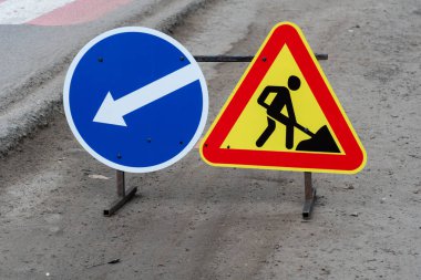 Bir şehir caddesine iki yol işareti yerleştirilmiş, biri solu gösteriyor ve diğeri de ileride inşaat çalışması olacağı uyarısını yapıyor.