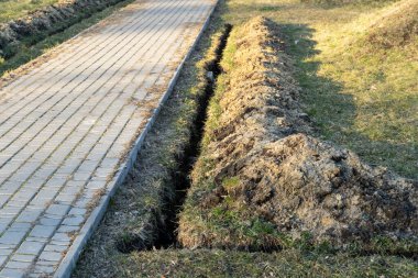 Kazı çalışmaları, öğleden sonra güneşli bir parkta çamur ve çimlerin rahatsız edildiği asfalt kaldırım kenarında yapılıyor.