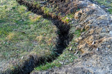 Toprağa kazılmış bir siper, açık toprak katmanlarını gösteriyor. Güneşli yeşil bir arazide doğa ve inşaatı harmanlıyor.