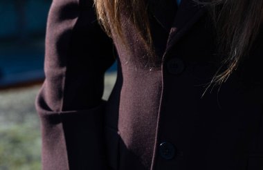 Koyu renk bir ceket giymiş, güneş ışığı saçına yansıyor ve parkın yakınındaki kumaşın dokusunu renklendiriyor.