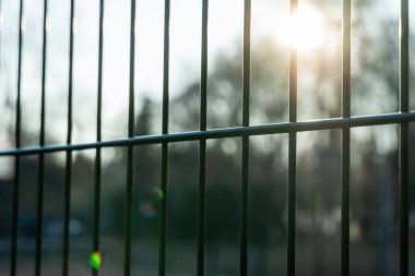 Metal bir çitin parmaklıklarından geçen güneş ışığı sabahın erken saatlerinde sakin bir parkta yumuşak gölgeler bırakır.