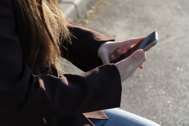 Bir kadın, aydınlık bir günde güneş ışığının sıcaklığının tadını çıkarırken akıllı telefonunu kontrol eder.