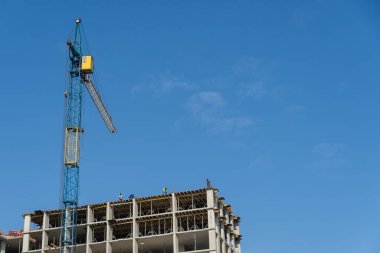 İnşaat sahasında, üzerinde büyük bir vinç bulunan bir yapı iskeleti üzerinde çalışanlar yer alıyor. Parlak mavi gökyüzü açık bir zemin sağlar