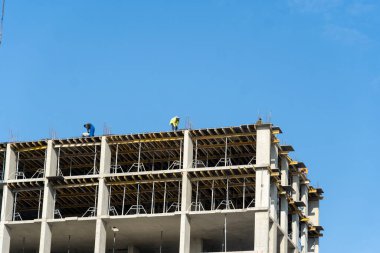 Güneşli bir günde, parlak mavi gökyüzü ile çevrili yüksek bir binanın çatısına iskelet döşeyen iki inşaat işçisi görülüyor.