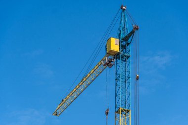 Büyük bir vinç parlak mavi gökyüzüne karşı dimdik ayakta duruyor, sarı ve mavi yapısı gün ışığında parlak güneş ışığı altında açıkça görülebiliyor.