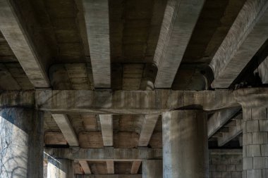 Büyük bir karayolu köprüsünün altında, beton sütunlar ve kirişler doğal ışıkla aydınlatılmış çarpıcı bir kentsel manzara yaratıyor, yapısal tasarım sergiliyor..