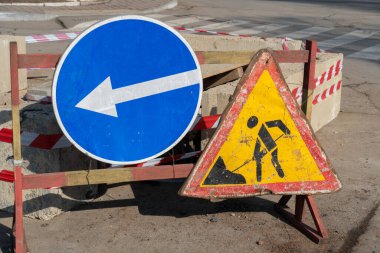 Bir cadde boyunca iki trafik levhası duruyor, biri sola doğru gidiyor ve diğeri inşaat işiyle ilgili uyarıda bulunuyor. Parlak güneş ışığı sahneyi aydınlatır