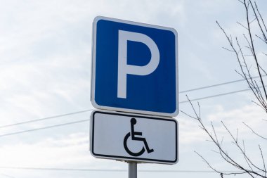 Mavi bir park yeri tabelası kentsel bir çevrede ulaşılabilir bir park yerini gösteriyor. Gökyüzü bulutlu ve ağaçlar yapraksız duruyor. Bu da ilkbaharın erken olduğunu gösteriyor.