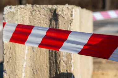 Kırmızı ve beyaz bant, güneşli bir günde açık havada beton bir bloğun etrafında bariyer oluşturan bir inşaat alanını işaretlemek için kullanılır.