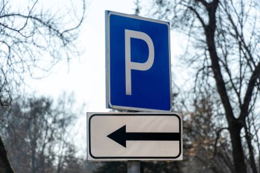 Güneşli bir günde, parkın ortasında çıplak ağaçlarla çevrili, solu gösteren bir tabelanın yanında mavi bir park işareti duruyor.