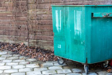 Etrafı dağınık kahverengi yapraklarla çevrili, ahşap bir çitin yanında yeşil bir çöp konteynırı duruyor.