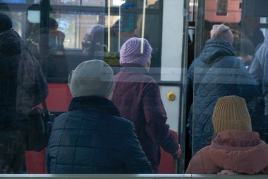 İki yaşlı insan otobüs durağında oturup, yoldan geçenleri izlerken, diğerleri soğuk hava koşullarında yoğun bir şehir ortamında otobüse biniyor.