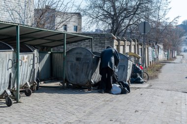 A person searches for items in large trash bins behind a building in an urban neighborhood under clear blue skies and bright sunlight clipart