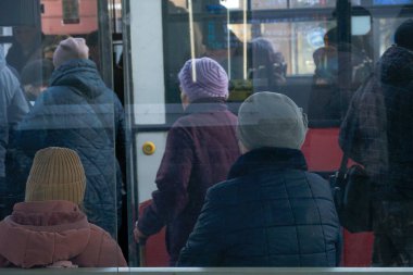 Yaşlı bireyler öğleden sonra yoğun bir otobüs durağında toplanarak diğerleri otobüse binerken toplu taşıma araçlarını bekliyorlar. Atmosfer canlı ve ortak hissettiriyor.