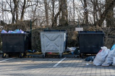 Etrafı dağınık çöp ve ağaçlarla çevrili bir otoparktaki çöplerle dolu. Bu da yakınlardaki atık yönetimi eksikliğini gösteriyor.