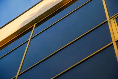 Pürüzsüz bir dış bina, parlak mavi gökyüzüne karşı batan güneşin sıcak renkleriyle aydınlatılan yansıtıcı cam paneller ve metal çerçeve içerir.