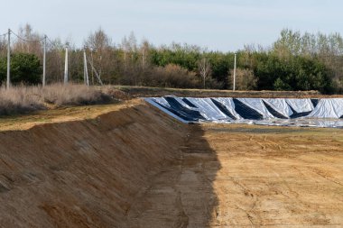 Çıplak ağaçlara ve elektrik direklerine bitişik, kırsal bir alanda siyah-beyaz koruyucu kaplamalı kazılmış bir çukur manzarası.