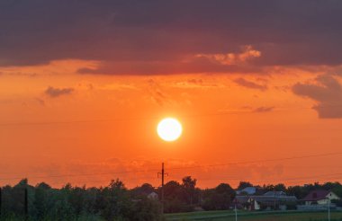 Canlı günbatımı, kırsal bir arazide turuncu bir parıltı oluşturur. Evlerin ve ağaçların siluetleri, gündüzleri geceye dönüşürken huzurlu bir atmosfer yaratıyor.