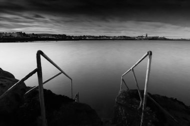 Çarpıcı bir siyah beyaz fotoğraf, doğal güzelliklere daldırılmış huzurlu bir su kütlesine uzanan bir dizi merdiveni yakalar.
