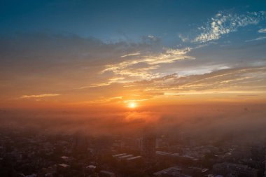 Gün batımında genişleyen bir şehri gösteren bir hava manzarası. Parlayan güneş gökyüzünde toplanan bulutların arasından güzel bir şekilde parlıyor.