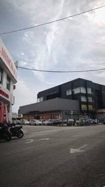 Bulutlu gökyüzü olan bir sokak sahnesi. Büyük pencereli siyah bir bina arka plandaki daha açık renkli binalara karşı duruyor. Arabalar cadde boyunca park halinde ve ön planda bir motosiklet görünüyor..