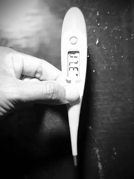 Siyah ve beyaz bir dijital termometre, koyu renkli bir yüzeyde tutuluyor. Termometrenin görüntüsü 37.7 derece gösteriyor..