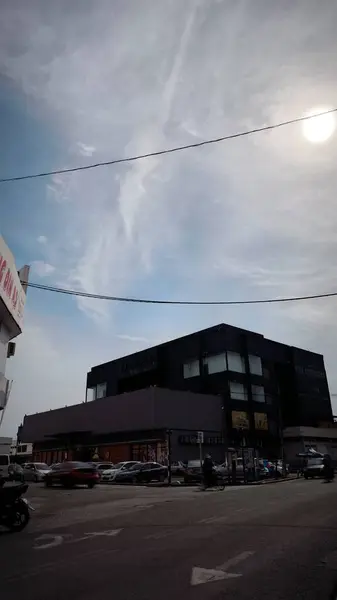 Bulutlu gökyüzü olan bir sokak sahnesi. Büyük pencereli siyah bir bina arka plandaki daha açık renkli binalara karşı duruyor. Arabalar cadde boyunca park halinde ve ön planda bir motosiklet görünüyor..