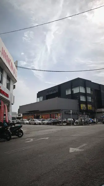 Bulutlu gökyüzü olan bir sokak sahnesi. Büyük pencereli siyah bir bina arka plandaki daha açık renkli binalara karşı duruyor. Arabalar cadde boyunca park halinde ve ön planda bir motosiklet görünüyor..