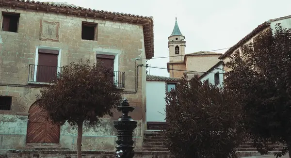 Büyüleyici köy meydanında tarihi taş binalar ve arka planda önemli bir kilise kulesi sergileniyor. Sahnede çıplak ağaçlar, antik bir su çeşmesi ve bir Avrupa köyünün zamansız özünü yakalayan geleneksel mimari yer alıyor..