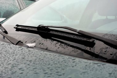 Yağmur damlaları ve silecekli bir arabanın ön camının yakın çekimi yağmurlu bir hava durumunu ve ıslak camları gösteriyor..