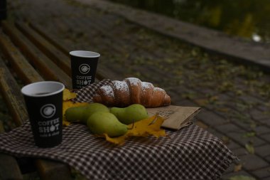 Yeşil armut, iki kahve fincanı, bir kruvasan ve parktaki ahşap bir bankta sarı yapraklar olan samimi bir sonbahar sahnesi. Sonbahar rahatlama ve ikramları için mükemmel bir mevsimlik atmosfer..