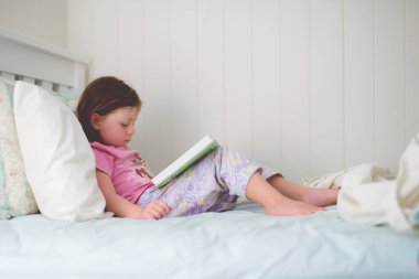 Genç bir kız pijamalarıyla yatağında oturmuş, rahat ve hafif bir yatak odasında kitap okuyor.