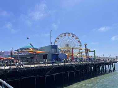 Kaliforniya 'daki ikonik Santa Monica İskelesi' nin canlı bir fotoğrafı. İçinde büyük bir dönme dolap, renkli lunapark trenleri ve güneşli bir günün keyfini çıkaran ziyaretçilerle dolu canlı bir sahil yolu var..