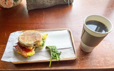 Taze ıspanak ve domatesle süslenmiş İngiliz çöreğinin üzerinde lezzetli bir yumurta ve pastırmalı sandviçin yer aldığı sıcak bir kahvaltı sahnesi, ahşap tepside bir fincan sade kahveyle paket servis ediliyor.