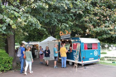 Barselona, İspanya - 21 Ağustos 2024: Bereketli yeşil bir parktaki canlı bir açık hava piyasa sahnesi, retro tarzı mavi gıda kamyonu krep servis ediyor. Barselona 'da. İspanya.