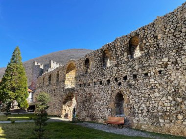 Açık mavi gökyüzü ve ormanlık bir tepenin zeminine karşı kurulmuş antik bir taş kalenin kalıntıları. Yıpranmış taş duvarlar, kemerli pencereler ve yarılmış kuleler ortaçağ mimarisini gözler önüne seriyor.