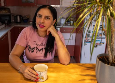 Samimi bir mutfakta elinde dekoratif bir kahve fincanı tutan düşünceli bir kadının portresi. Sahne, ev bitkileriyle ve seyahat mıknatıslarıyla dolu bir buzdolabıyla sıcak bir şekilde aydınlatıldı..