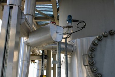 Endüstriyel metalik boruların yakın görüntüsü ve modern bir tesiste basınç sensörü. Görüntü temiz mühendislik yapılarını, cıvataları ve çelik çerçevenin altında sağlam bir mekanik ortamı vurguluyor..