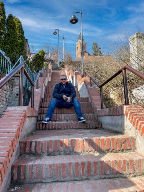 Güneş gözlüklü ve siyah ceketli bir adam dışarıdaki kırmızı tuğla merdivenlerde poz veriyor. Olay yeri eski tip sokak lambaları, çıplak ağaçlar ve berrak mavi gökyüzünün altında kırsal tuğla bir kule ile çerçevelenmiş. Manzaralı bir şehirde sakin bir anı yakalıyor..