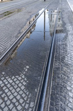 Şehir tramvay izleri, kaldırım taşı caddesinde yansıtıcı bir su birikintisi olan tarihi binaların aynalı görüntülerini yakalıyor. Yağmurdan sonra çekilen sahne, sakin bir şehirde simetri, doku ve kentsel altyapıyı vurguluyor..