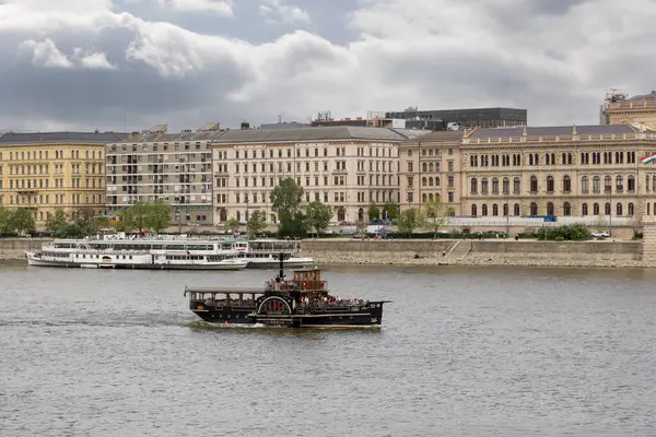 Budapeşte, Macaristan - 01 Mayıs 2025: Macaristan 'ın Budapeşte kentindeki klasik Avrupa binalarının zarif zeminine karşı Tuna Nehri boyunca yolcularla birlikte eski tarz bir gezi teknesi denize açılıyor.
