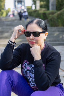 Kendine güveni tam bir kadın, dışarıda taş basamaklarda oturuyor, güneş gözlüğünü ayarlarken doğal güneş ışığı altında gülümsüyor, kendine güvenini ve rahat şehir yaşam tarzını ifade ediyor..