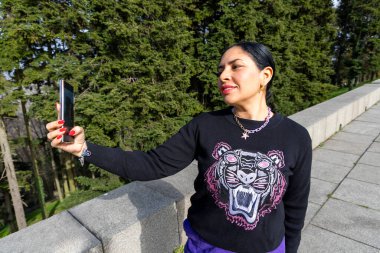 Parkta akıllı telefonuyla fotoğraf çekerken gülümseyen şık bir kadın cesur bir kaplan süveteri giyiyor ve renkli mücevherlerle süslenmiş neşeli bir anı yakalıyor.