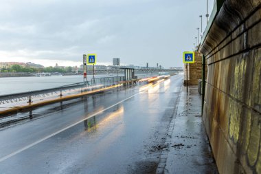 Yağmurlu nehir kenarı yolu. Arabalardan gelen bulanık ışık yolları, yaya geçidi işaretleri ve ıslak kaldırıma yansıyan bulutlu gökyüzü.