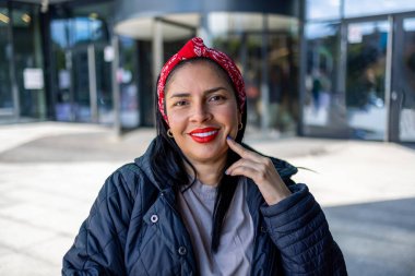 Uzun siyah saçlı, kırmızı rujlu, kırmızı bandana takan, koyu renk ceketli, modern şehir ortamında açık havada duran gülümseyen bir kadının portresi. Görüntü sıcaklığı, güveni ve günlük tarzı yakalar..