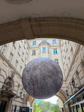 Tarihi binalar arasında asılı duran büyük, gerçekçi bir ay sanat tesisi. Sürrealite sahnesi, klasik Avrupa mimarisi ile çağdaş sanata zıt düşüyor.