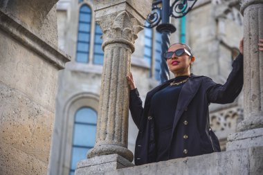 Kendine güvenen kadın, tarihi mimarinin çerçevelediği süslü taş sütunlar arasında poz veriyor. Zarif bir ortam ve cesur moda Avrupa cazibesiyle çarpıcı bir seyahat ve moda portresi oluşturur..