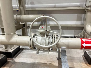 Endüstriyel boru sisteminin yüksek çözünürlüklü görüntüsü. El tekerleği olan büyük bir manuel vana içeriyor. Kurulum kapalı, bej boyalı borular ve kırmızı yönlü akış göstergesi ile.