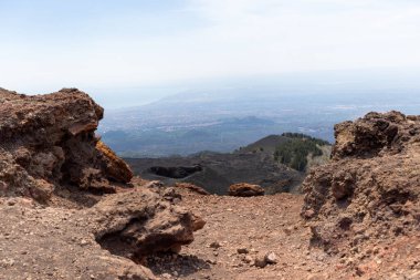 Kıyı şeridi panoramik manzaralı bir dağ zirvesi üzerinde dramatik volkanik arazi. Kayalık, kırmızımsı-kahverengi ön plan, bereketli vadi ve uzaktaki ışıldayan denizle çelişiyor..