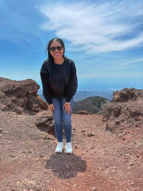 Dağılmış bulutlarla dolu parlak mavi gökyüzünün altında kayalık bir dağın tepesinde gülümseyen kadın. Arka plan uzak manzara görüntülerini gözler önüne seriyor, macera, özgürlük ve seyahat çağrıştırıyor.